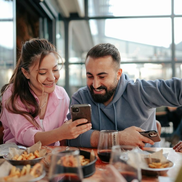 Zwei Personen sitzen an einem Tisch, lächeln und schauen auf ein Smartphone. Essen und Getränke sind sichtbar.